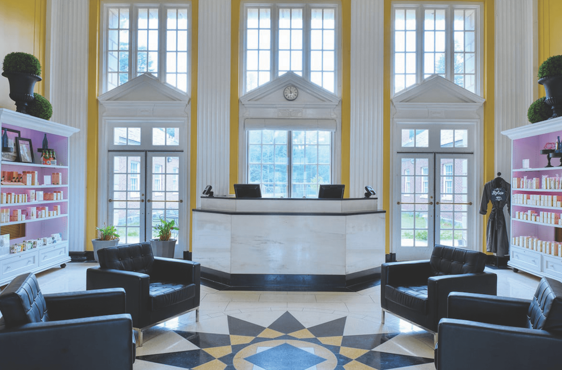 A spacious, elegant lobby with tall windows, a white reception desk, black leather chairs, geometric tile flooring, and shelves displaying wellness products on both sides. Natural light fills the room, creating an atmosphere of relaxation.
